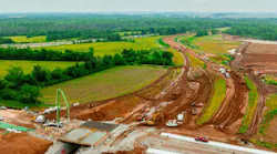An aerial view of construction on the MetroLink extension to MidAmerica St. Louis Airport shows graded dirt roadbed, a concrete underpass taking shape and heavy equipment surrounded by farmland and trees.