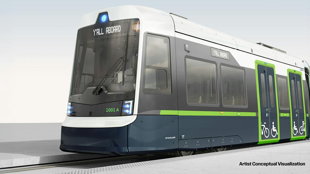 An artist's conceptual rendering of a Stadler light-rail vehicle in navy, gray and white with green accents, showing a 'Y'ALL ABOARD' destination sign, vehicle number 1001 A and bike and wheelchair accessibility symbols at the doors.