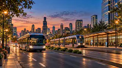 An illustrated city street at dusk shows a white bus and a multi-car light rail vehicle with glowing headlights, a lit station platform with waiting passengers and a skyline of lit skyscrapers under an orange and purple sky.