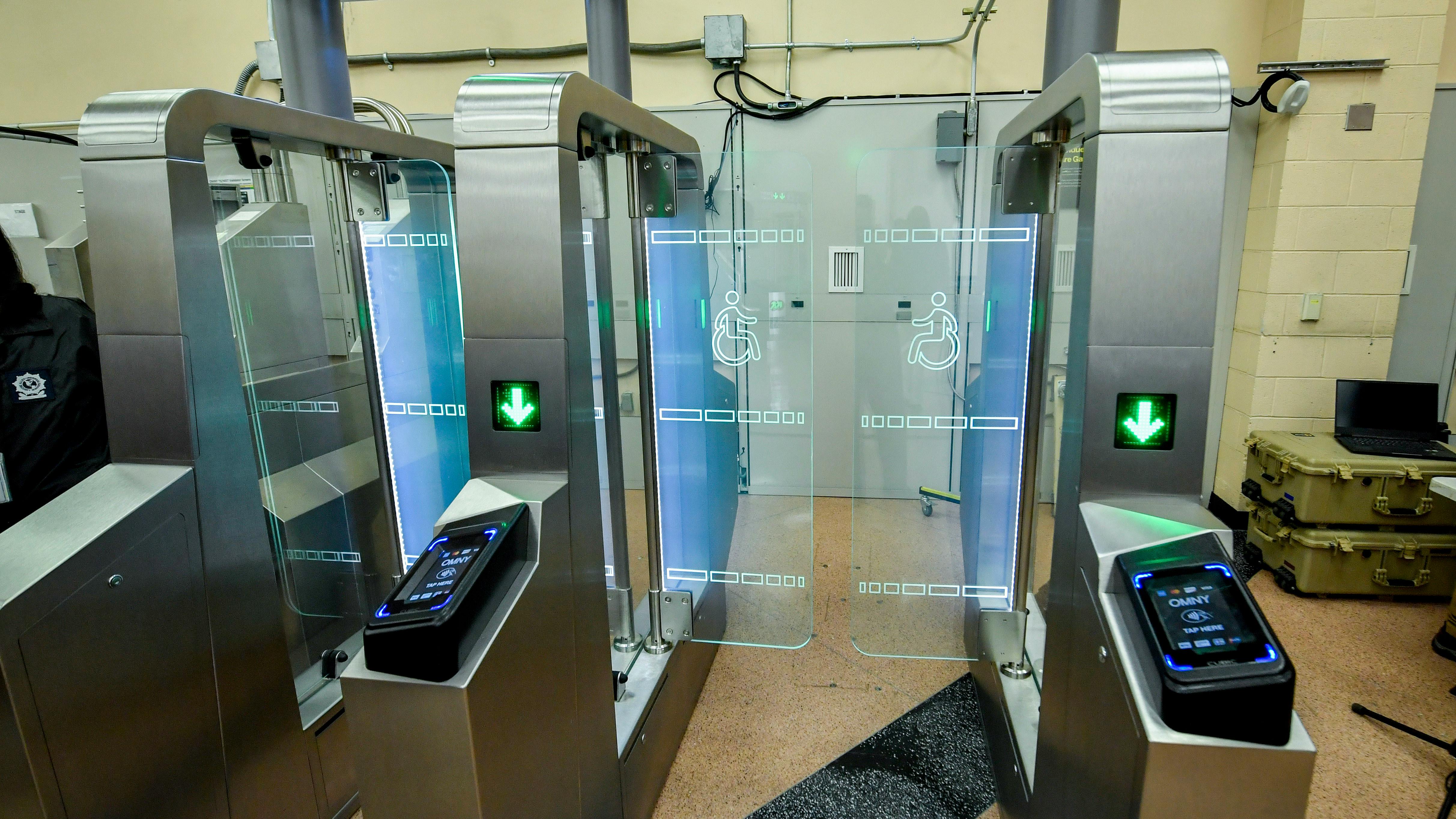 A pair of stainless steel fare gates with glass panels, green arrow indicators and wheelchair-accessibility symbols stands in an indoor space, with OMNY 'Tap Here' readers mounted on the gate pedestals.