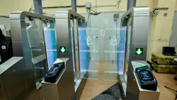 A pair of stainless steel fare gates with glass panels, green arrow indicators and wheelchair-accessibility symbols stands in an indoor space, with OMNY 'Tap Here' readers mounted on the gate pedestals.