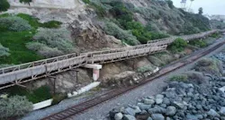 An elevated wooden and metal walkway runs along a hillside above a rail line, with a concrete support pier, rocks on the near side and coastal vegetation on the slope.