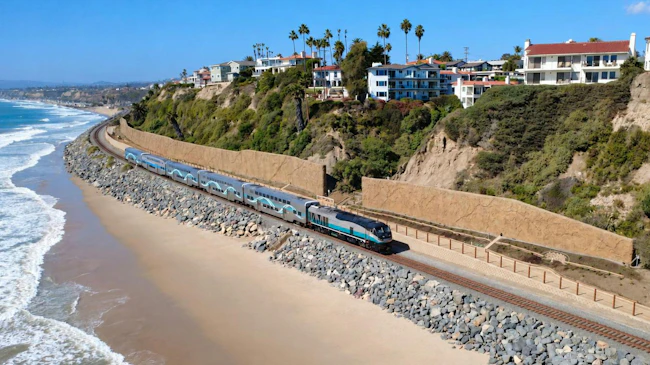A blue, gray and white Metrolink commuter train travels along a rock-armored coastal rail line beside a sandy beach and ocean surf, with hillside homes and tall retaining walls above.