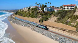 A blue, gray and white Metrolink commuter train travels along a rock-armored coastal rail line beside a sandy beach and ocean surf, with hillside homes and tall retaining walls above.