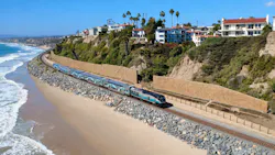 A blue, gray and white Metrolink commuter train travels along a rock-armored coastal rail line beside a sandy beach and ocean surf, with hillside homes and tall retaining walls above.