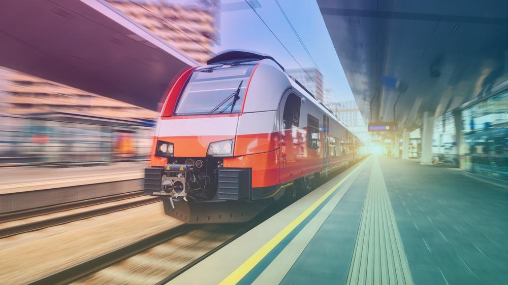 A red, white and gray electric passenger train speeds into a station with motion blur, passing a platform with a yellow safety line and tactile paving under a stylized teal and purple color grade.