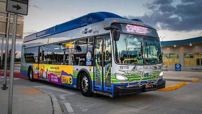 A Miami-Dade County transit bus, unit 19118, with blue, green and white livery and a Spanish-language radio advertisement on its side, is parked at a curb near a terminal building at dusk.
