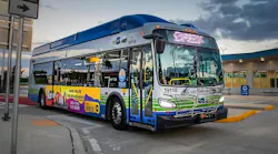 A Miami-Dade County transit bus, unit 19118, with blue, green and white livery and a Spanish-language radio advertisement on its side, is parked at a curb near a terminal building at dusk.