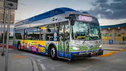 A Miami-Dade County transit bus, unit 19118, with blue, green and white livery and a Spanish-language radio advertisement on its side, is parked at a curb near a terminal building at dusk.