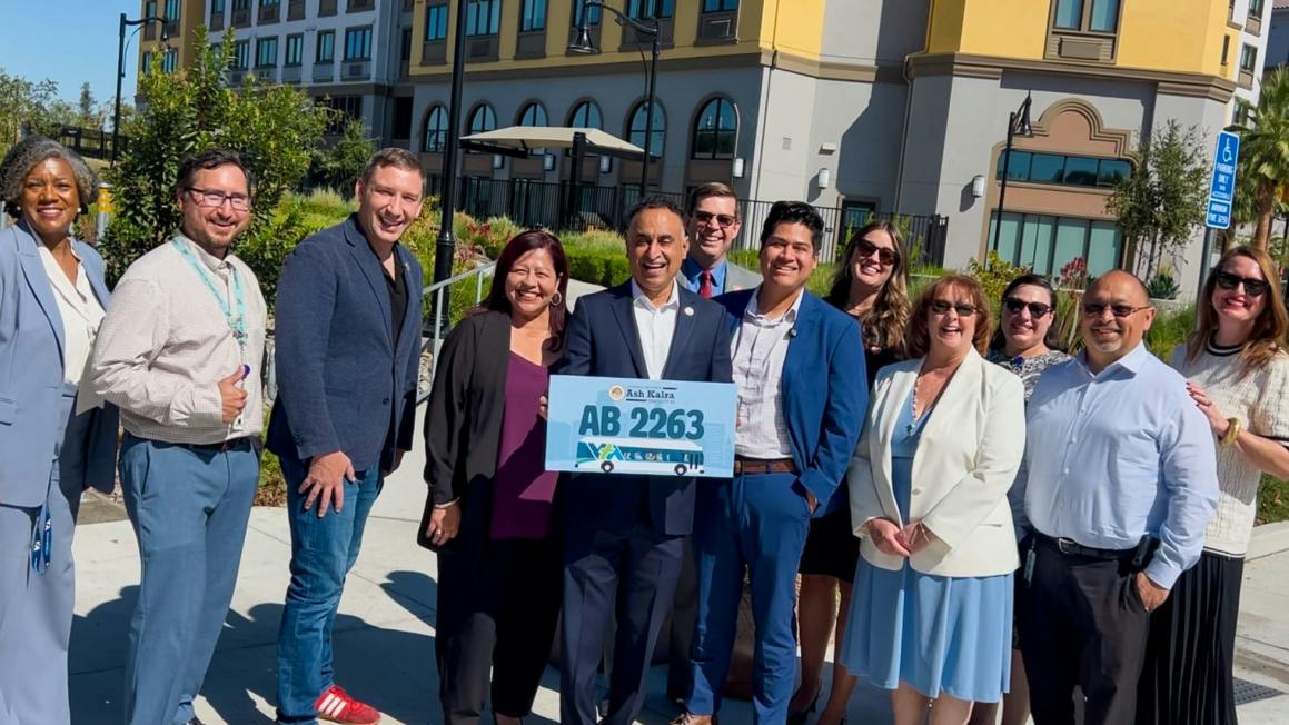 A group of eleven people stand outdoors smiling, with a man in the center holding a sign reading &lsquo;Ash Kalra, AB 2263&rsquo; featuring a bus graphic.