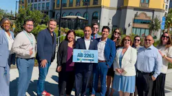 A group of eleven people stand outdoors smiling, with a man in the center holding a sign reading ‘Ash Kalra, AB 2263’ featuring a bus graphic.