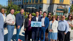 A group of eleven people stand outdoors smiling, with a man in the center holding a sign reading ‘Ash Kalra, AB 2263’ featuring a bus graphic.