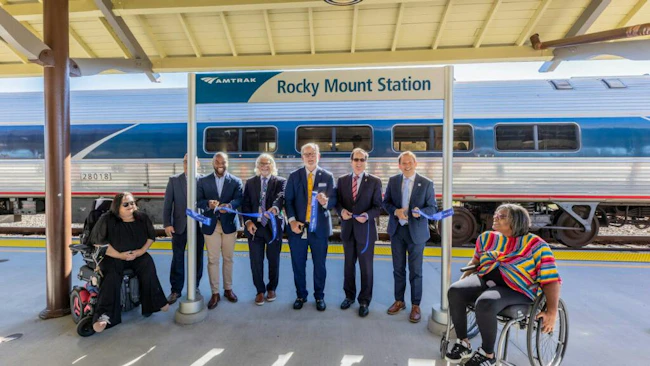 Officials and community members hold a blue ribbon at a ribbon-cutting ceremony under the platform canopy at Rocky Mount Station, with an Amtrak train stopped behind them and two people seated in wheelchairs at either end of the group.