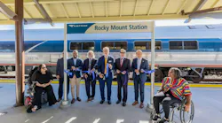 Officials and community members hold a blue ribbon at a ribbon-cutting ceremony under the platform canopy at Rocky Mount Station, with an Amtrak train stopped behind them and two people seated in wheelchairs at either end of the group.