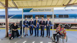 Officials and community members hold a blue ribbon at a ribbon-cutting ceremony under the platform canopy at Rocky Mount Station, with an Amtrak train stopped behind them and two people seated in wheelchairs at either end of the group.