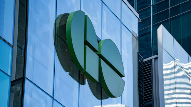 A large green sculptural logo, made of split circular and rectangular segments, is mounted on a blue glass building facade, with neighboring glass towers in the background.