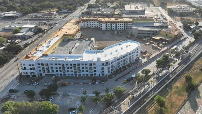 An aerial view of a multistory mixed-use building under construction beside a rail line and station platform with canopies, with a bus on the adjacent road, surrounding streets, parking lots and trees.