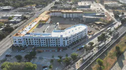An aerial view of a multistory mixed-use building under construction beside a rail line and station platform with canopies, with a bus on the adjacent road, surrounding streets, parking lots and trees.