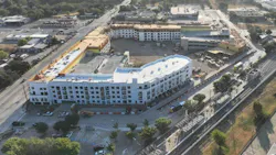An aerial view of a multistory mixed-use building under construction beside a rail line and station platform with canopies, with a bus on the adjacent road, surrounding streets, parking lots and trees.