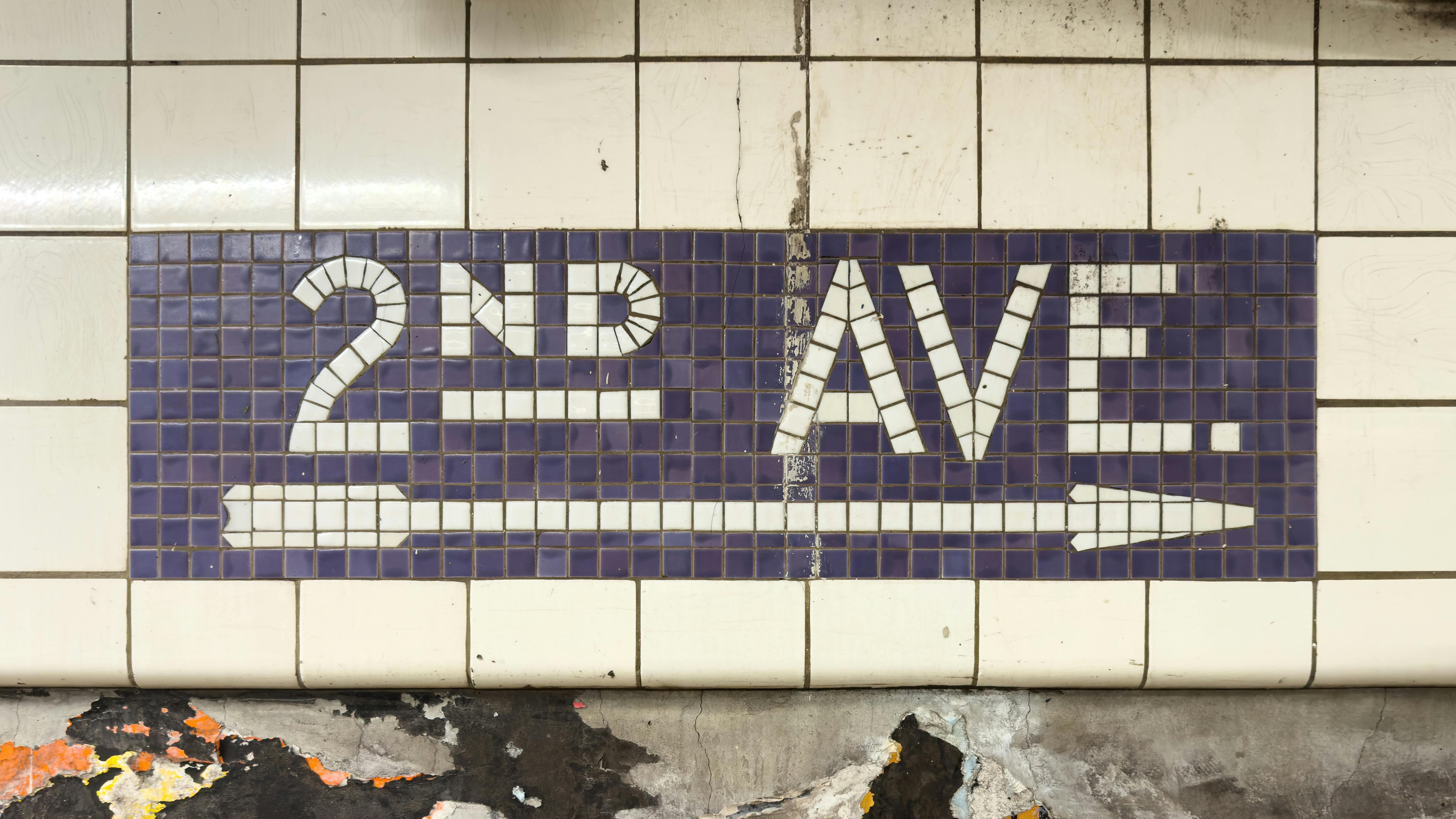 A worn subway station wall, with a mosaic sign reading &lsquo;2ND AVE.&rsquo; and an arrow in white tile on a purple background, above a tiled wall with peeling layers of old posters.