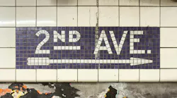 A worn subway station wall, with a mosaic sign reading ‘2ND AVE.’ and an arrow in white tile on a purple background, above a tiled wall with peeling layers of old posters.