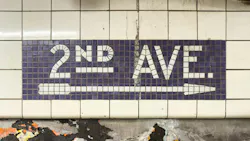 A worn subway station wall, with a mosaic sign reading ‘2ND AVE.’ and an arrow in white tile on a purple background, above a tiled wall with peeling layers of old posters.