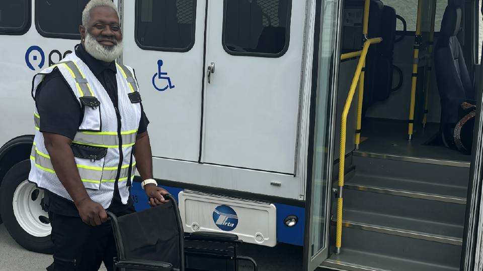A smiling transit worker in a high-visibility vest stands beside an empty wheelchair next to a paratransit-style bus with its door open, showing handrails, steps and a wheelchair accessibility symbol.