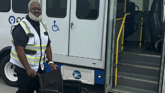 A smiling transit worker in a high-visibility vest stands beside an empty wheelchair next to a paratransit-style bus with its door open, showing handrails, steps and a wheelchair accessibility symbol.