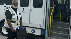 A smiling transit worker in a high-visibility vest stands beside an empty wheelchair next to a paratransit-style bus with its door open, showing handrails, steps and a wheelchair accessibility symbol.