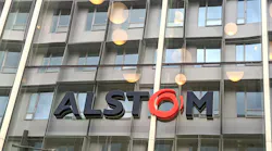 Close-up of the Alstom logo in dark gray and red lettering mounted on a glass office building facade, with round pendant lights visible through the windows.