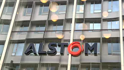 Close-up of the Alstom logo in dark gray and red lettering mounted on a glass office building facade, with round pendant lights visible through the windows.