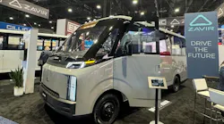 A silver Zavir electric shuttle bus on display at a trade show, parked next to a sign reading ‘ZAVIR, Drive the Future,’ with other exhibit booths in the background.