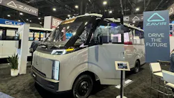 A silver Zavir electric shuttle bus on display at a trade show, parked next to a sign reading ‘ZAVIR, Drive the Future,’ with other exhibit booths in the background.