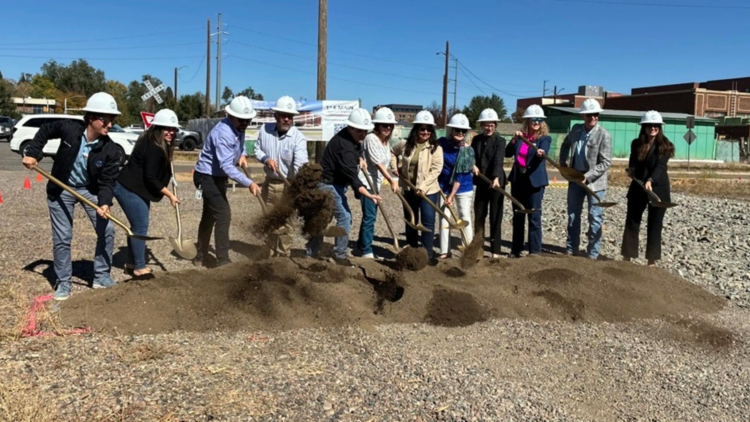 A line of eleven people in white hard hats shovel dirt with gold-colored shovels at a groundbreaking ceremony on a gravel lot, with a rendering banner of the transit center, a railroad crossing sign and brick buildings behind them.