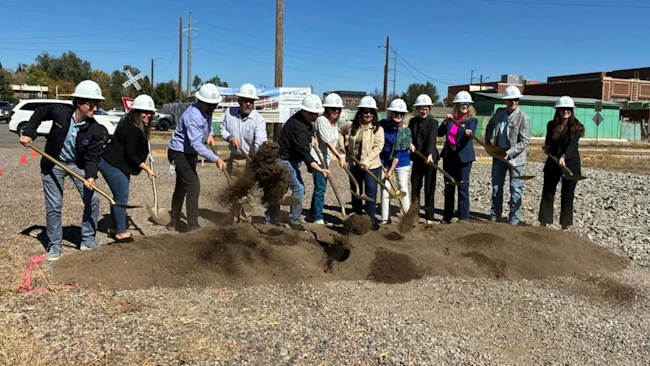A line of eleven people in white hard hats shovel dirt with gold-colored shovels at a groundbreaking ceremony on a gravel lot, with a rendering banner of the transit center, a railroad crossing sign and brick buildings behind them.