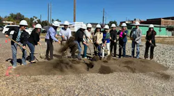 A line of eleven people in white hard hats shovel dirt with gold-colored shovels at a groundbreaking ceremony on a gravel lot, with a rendering banner of the transit center, a railroad crossing sign and brick buildings behind them.