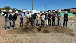 A line of eleven people in white hard hats shovel dirt with gold-colored shovels at a groundbreaking ceremony on a gravel lot, with a rendering banner of the transit center, a railroad crossing sign and brick buildings behind them.