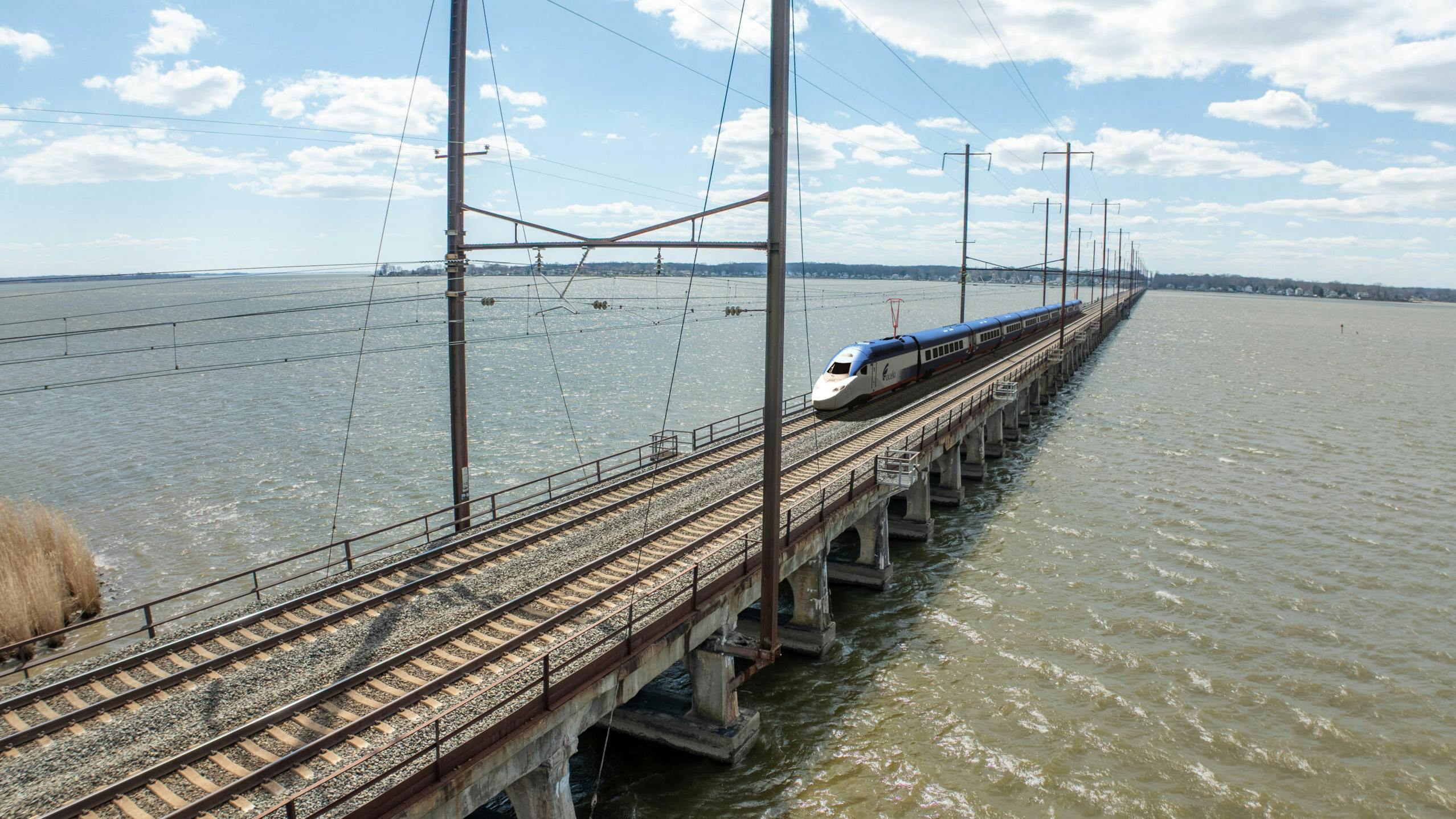 An Amtrak high-speed train crosses a long railway bridge over a wide body of water under a partly cloudy sky..
