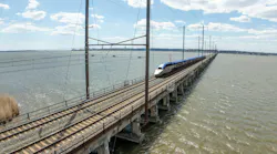 An Amtrak high-speed train crosses a long railway bridge over a wide body of water under a partly cloudy sky..
