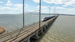 An Amtrak high-speed train crosses a long railway bridge over a wide body of water under a partly cloudy sky..