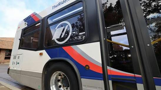 A close-up of the side of a white transit bus with blue and red stripes, a circular logo on the window and &ldquo;TheRide.org&rdquo; printed along the roofline, parked at a curb.