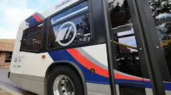 A close-up of the side of a white transit bus with blue and red stripes, a circular logo on the window and “TheRide.org” printed along the roofline, parked at a curb.