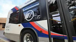 A close-up of the side of a white transit bus with blue and red stripes, a circular logo on the window and “TheRide.org” printed along the roofline, parked at a curb.