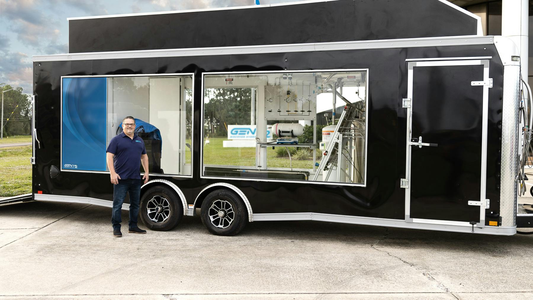 Chris Wallington stands beside a black enclosed trailer with large windows showing hydrogen fueling and testing equipment, including gauges, tubing and a gas cylinder, with a GEN H2 logo on the side.