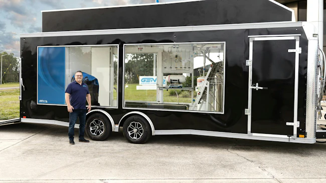 Chris Wallington stands beside a black enclosed trailer with large windows showing hydrogen fueling and testing equipment, including gauges, tubing and a gas cylinder, with a GEN H2 logo on the side.