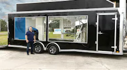 Chris Wallington stands beside a black enclosed trailer with large windows showing hydrogen fueling and testing equipment, including gauges, tubing and a gas cylinder, with a GEN H2 logo on the side.