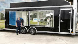 Chris Wallington stands beside a black enclosed trailer with large windows showing hydrogen fueling and testing equipment, including gauges, tubing and a gas cylinder, with a GEN H2 logo on the side.