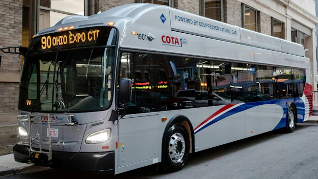 A silver, red and blue COTA bus, unit 1901, labeled &ldquo;Powered by Compressed Natural Gas,&rdquo; displays &ldquo;90 Ohio Expo Ctr&rdquo; on its destination sign while parked at a curb beside a brick building.