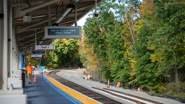 A train station platform with a sign reading &lsquo;Track 1&rsquo; and an electronic display showing &lsquo;Train No. 56 to St. Albans, Status - Delayed,&rsquo; with a worker in an orange vest on the platform and autumn trees along the tracks.