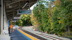A train station platform with a sign reading ‘Track 1’ and an electronic display showing ‘Train No. 56 to St. Albans, Status - Delayed,’ with a worker in an orange vest on the platform and autumn trees along the tracks.